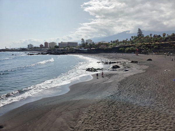 Vista panorámica de la extensa playa de arena volcánica de Punta Brava con el Teide semiencubierto por nubes al fondo.